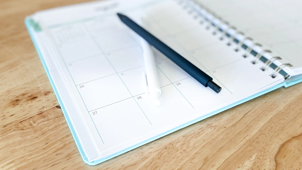 An open monthly planner with two pens resting on its calendar pages on a wooden desk